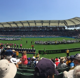 A large sports stadium filled with spectators, a well-maintained green field in the center, and a group of people forming lines as they prepare for an event. The seating areas are crowded, and the atmosphere appears lively with fans wearing various team colors and merchandise. Overhead, clear skies and bright sunlight illuminate the venue.