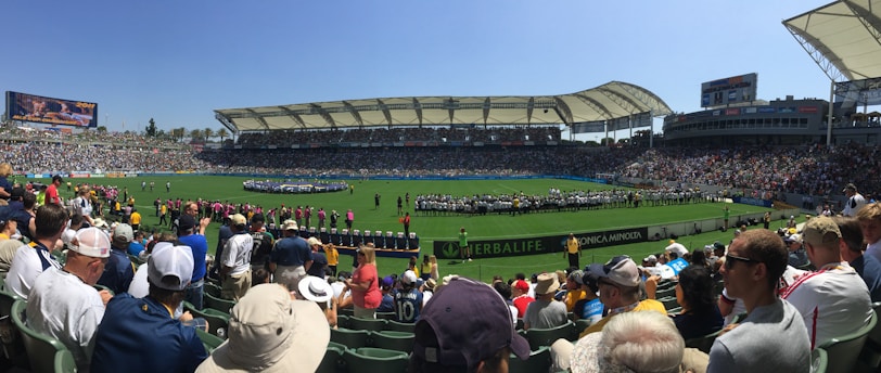 A large sports stadium filled with spectators, a well-maintained green field in the center, and a group of people forming lines as they prepare for an event. The seating areas are crowded, and the atmosphere appears lively with fans wearing various team colors and merchandise. Overhead, clear skies and bright sunlight illuminate the venue.