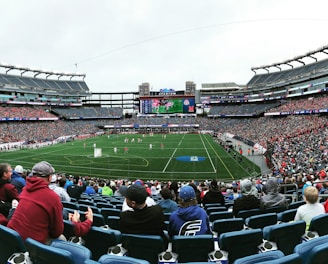 A large sports stadium filled with spectators. The field is green with white markings, and players in red and white uniforms are actively engaged in a game. The stands are packed with people wearing various colored clothing, and some sections of the crowd are more densely populated. Digital screens and advertisements are visible around the stadium, adding to the vibrant atmosphere.