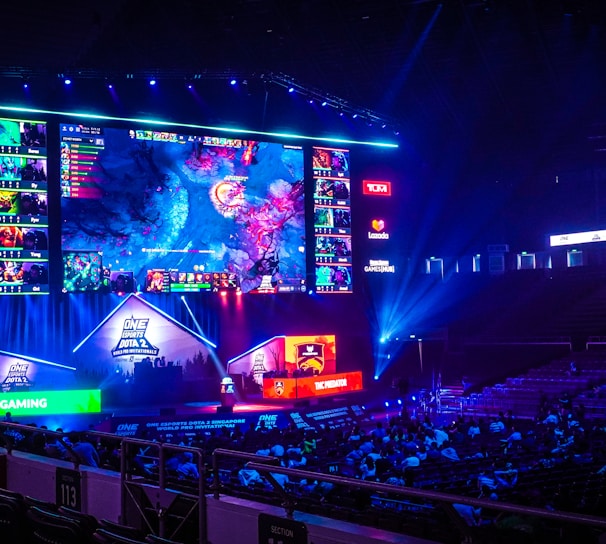 A large esports arena featuring a massive screen displaying a video game, surrounded by sponsor logos and team names. The auditorium is filled with spectators watching the gaming event. Neon blue and purple lights illuminate the stage, creating a vibrant atmosphere.
