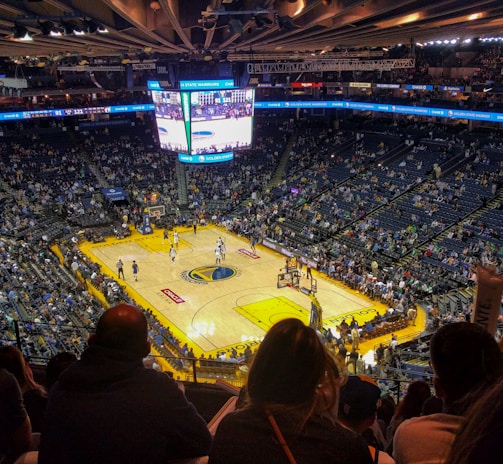 An indoor basketball arena filled with spectators. The court features a central logo and is surrounded by partially occupied tiered seating. A large scoreboard hangs above the center of the court, displaying the game score and time. Ambient lighting highlights the players on the court and the vibrant atmosphere of a live sports event.