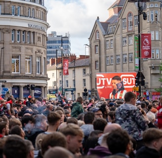 A large crowd of people, many wearing red and white, gather in a city square. They are watching an event broadcast on a large screen, which displays a man speaking into a microphone with logos suggesting a sporting event. Surrounding buildings are historical and commercial, with banners indicating a connection to the Premier League.