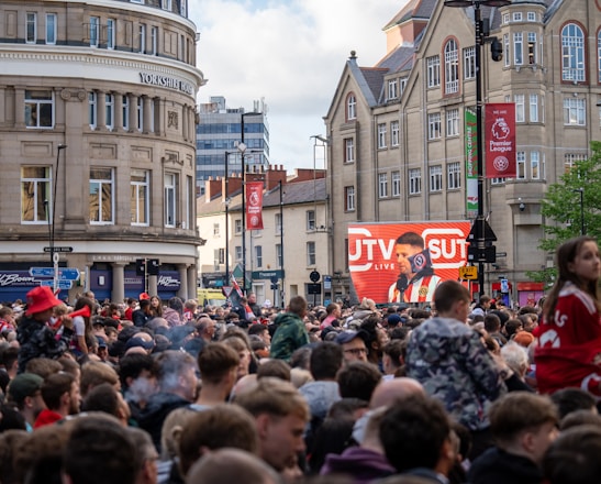A large crowd of people, many wearing red and white, gather in a city square. They are watching an event broadcast on a large screen, which displays a man speaking into a microphone with logos suggesting a sporting event. Surrounding buildings are historical and commercial, with banners indicating a connection to the Premier League.